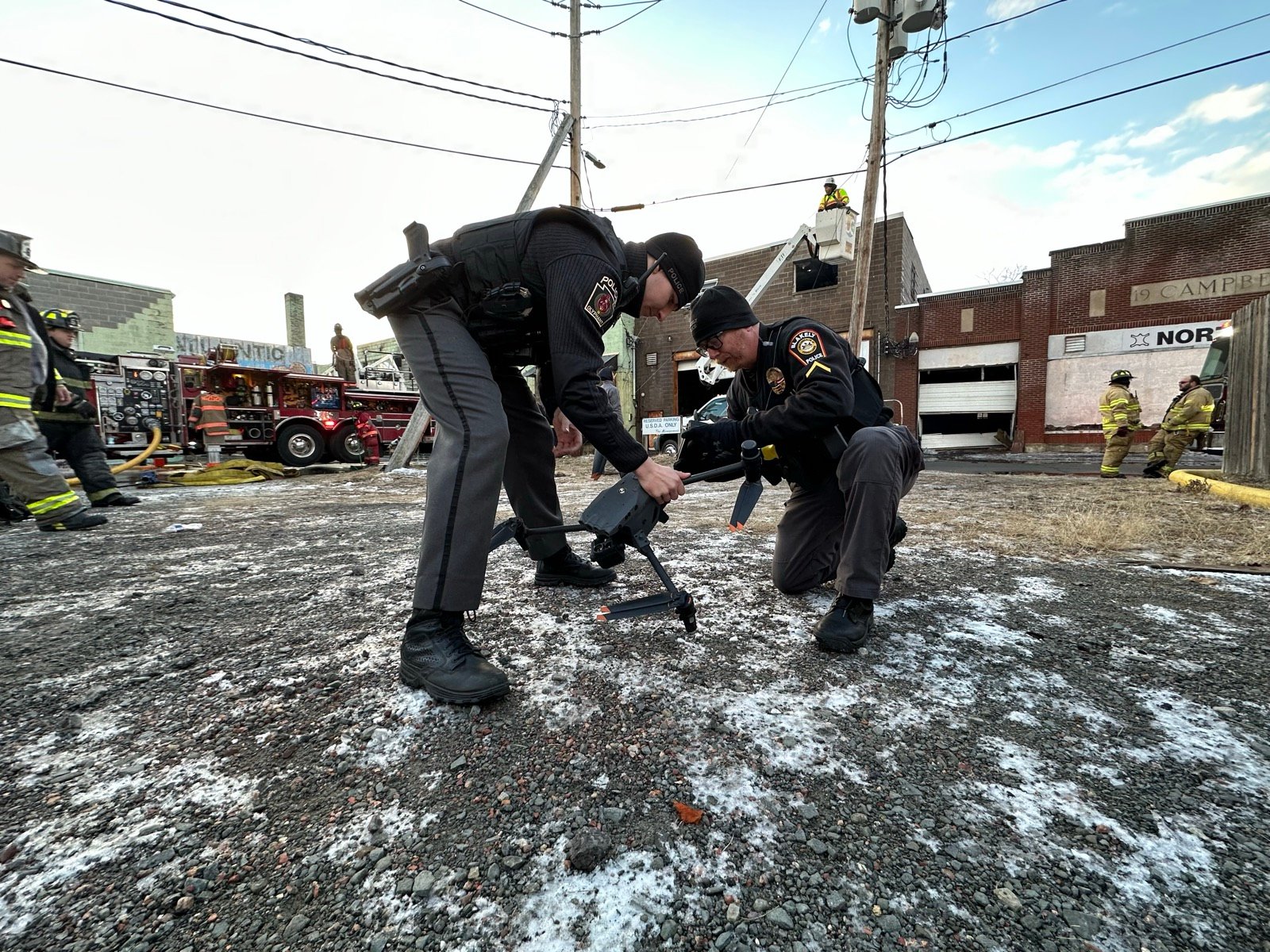 Two police officers setting up a drone at a fire scene as firefighters work behind them